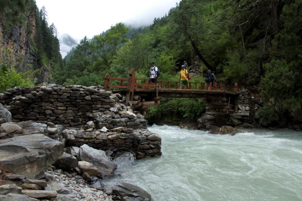 A scenic view of a wooden bridge in Nepal, crossing a turbulent river. Hikers in various attire traverse the bridge amidst a backdrop of towering cliffs, lush greenery, and low-hanging mist. A traditional stone structure stands near the bank, embodying the rustic charm of the region.