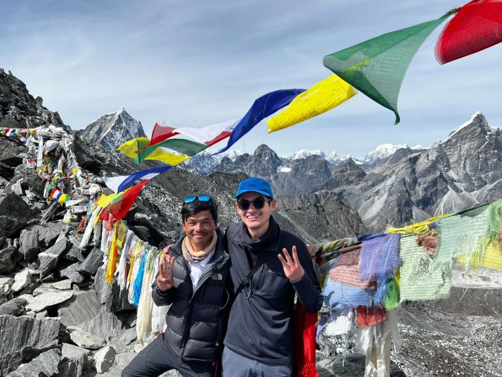 Guide Jiten and trekker Luke posing with peace signs on the Three Passes Trek in the Khumbu region, with colorful prayer flags and snow-capped mountains in the background.