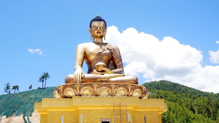 A large, golden Buddha statue seated in a meditative pose atop a platform against a backdrop of blue sky with scattered clouds, flanked by a verdant mountain landscape, characteristic of Bhutanese terrain.