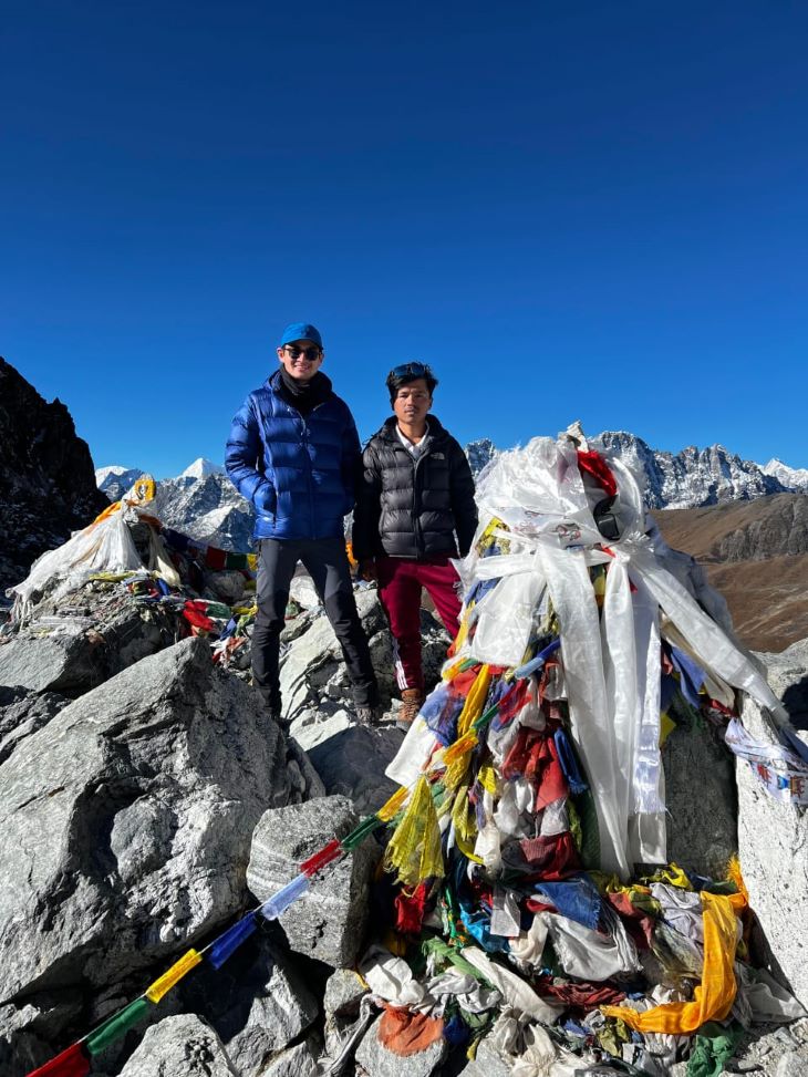 Congratulations to our guide Jiten and trekker Luke for their accomplishment on the Three Passes Trek, shown standing triumphantly amidst prayer flags at a high-altitude pass with the stark Himalayan range in the background.