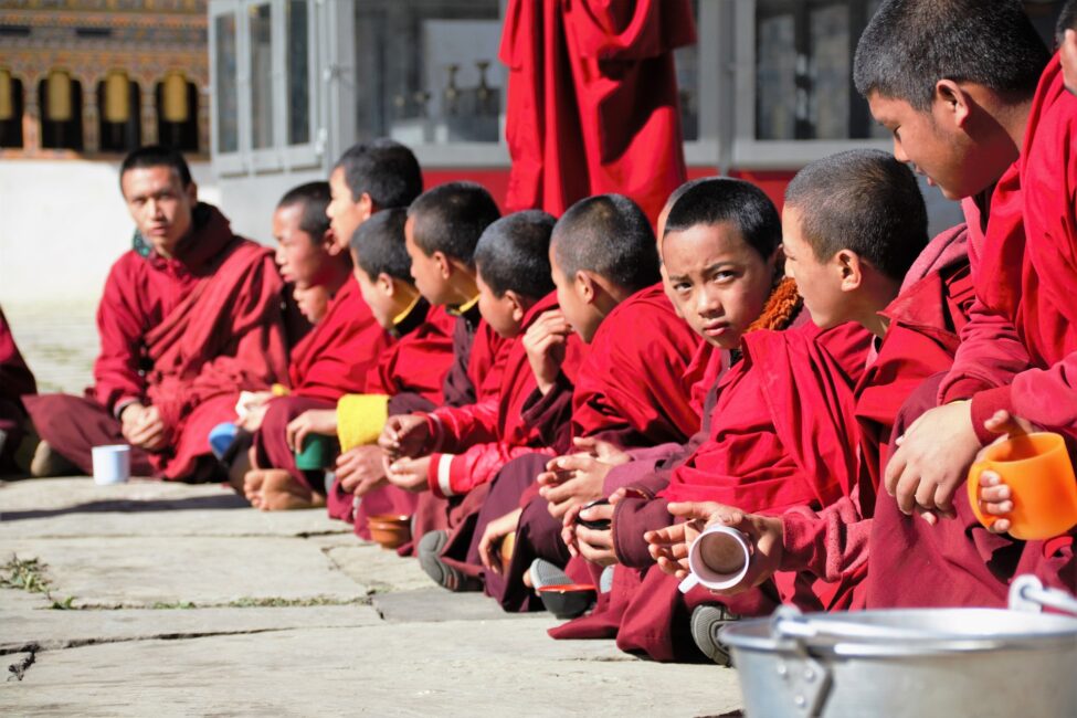 A group of young monks in traditional red robes sitting together on the ground, some with bowls in their hands, in a courtyard possibly within a monastery in Bhutan