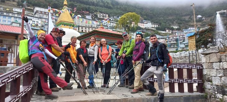 A diverse group of trekkers with bright smiles, geared up with backpacks and trekking poles, poses on a bridge in Namche Bazaar, with the vibrant buildings of the town and a prominent golden stupa in the background, ready to embark on their adventure towards Everest Base Camp.